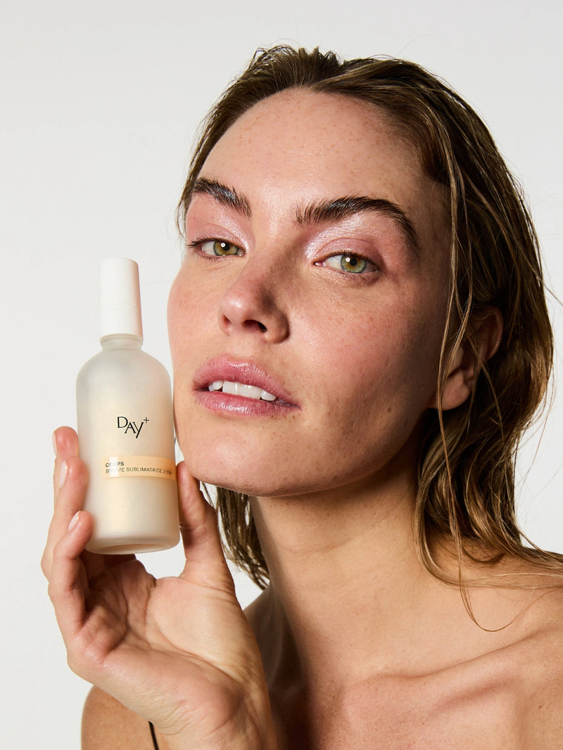 Brume Sublimatrice 3-en-1 dans les mains d'une femme au maquillage frais et lumineux et aux cheveux effet mouillé.