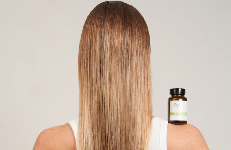 Pilulier de Gelules Cheveux Fortifiantes posé sur l'épaule d'une femme prise de dos avec de longs cheveux blond.