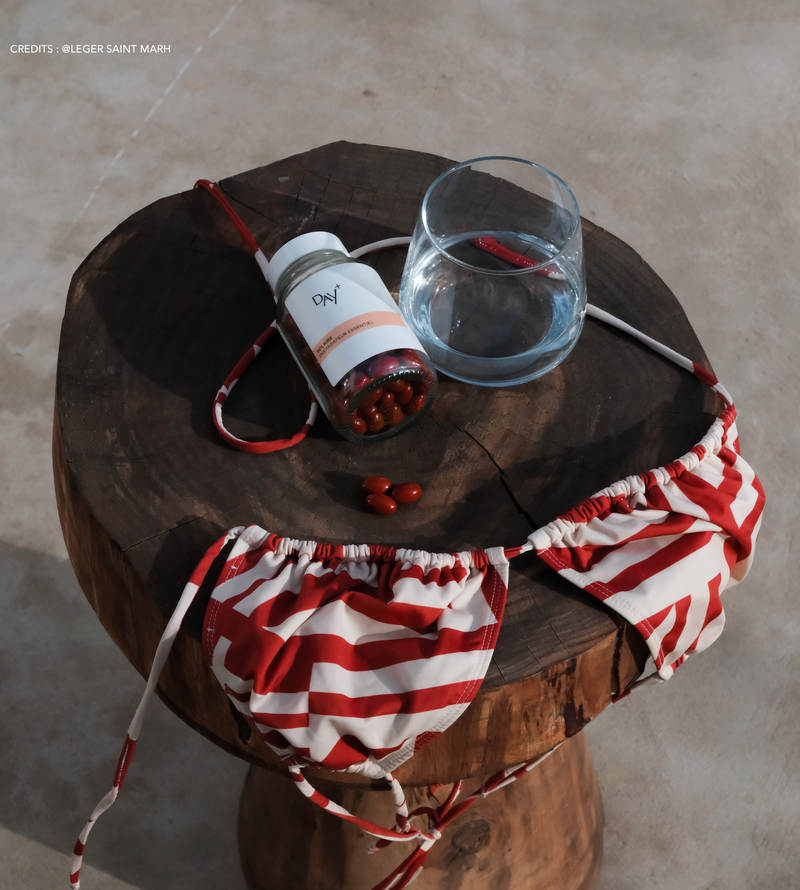 Le soin préparateur essentiel posé sur un tabouret en rondin de bois avec un verre d'eau et un haut de maillot de bain triangle rayé rouge et blanc