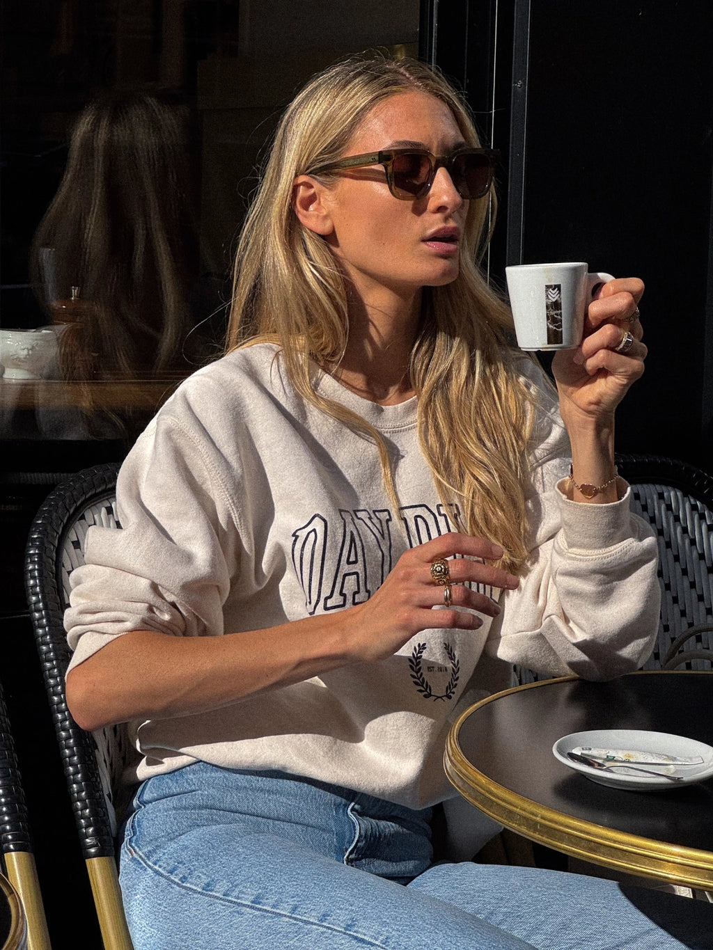 Femme assise à un café portant le sweatshirt beige de la collection capsule Day+ University.