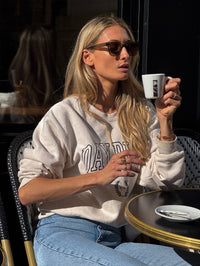 Femme assise à un café portant le sweatshirt beige de la collection capsule Day+ University.