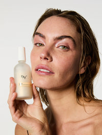 Brume Sublimatrice 3-en-1 dans les mains d'une femme au maquillage frais et lumineux et aux cheveux effet mouillé.