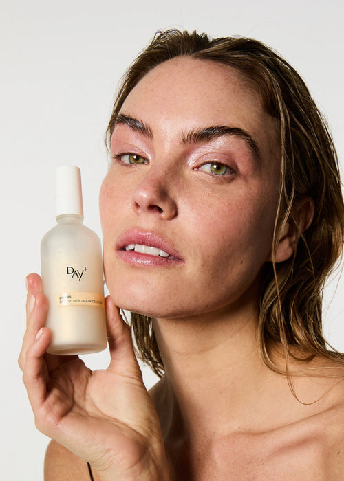 Brume Sublimatrice 3-en-1 dans les mains d'une femme au maquillage frais et lumineux et aux cheveux effet mouillé.