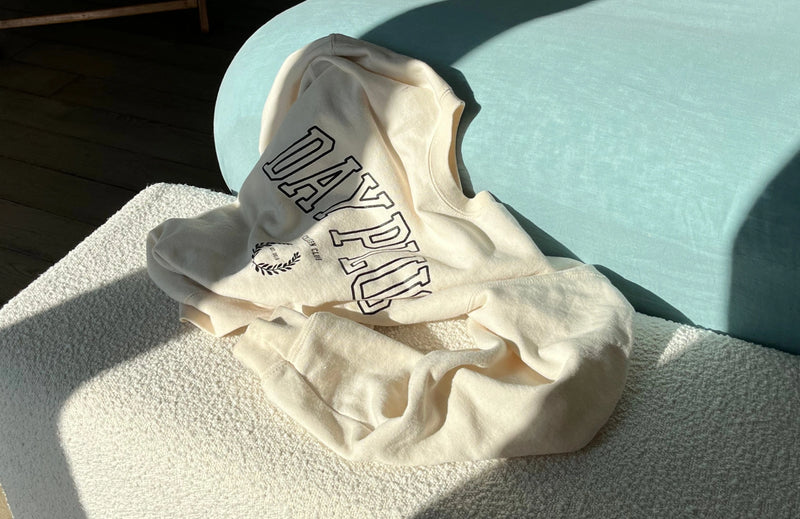 Sweatshirt beige avec logo noir de la collection capsule Day+ University posé sur un lit.