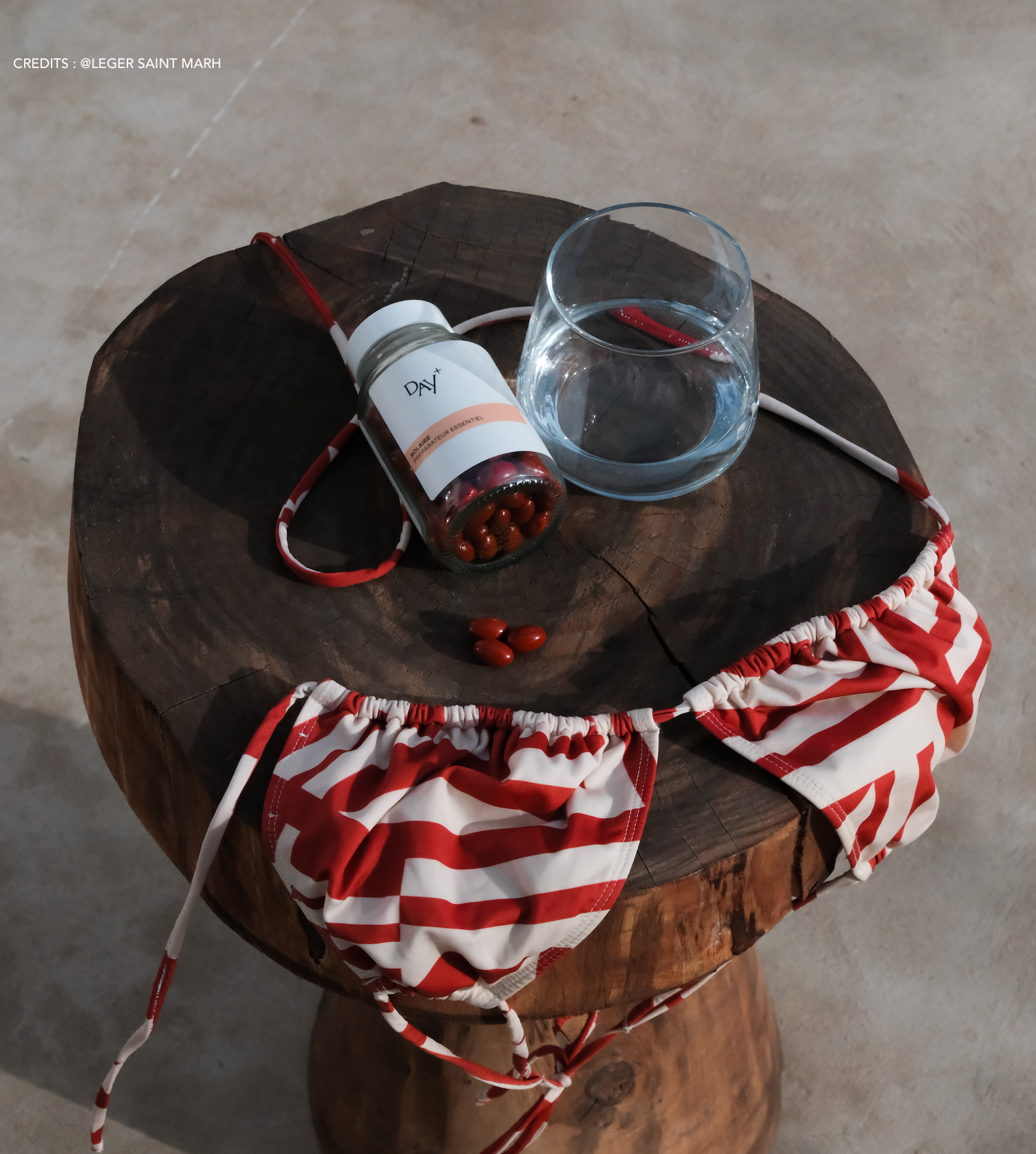 Le soin préparateur essentiel posé sur un tabouret en rondin de bois avec un verre d'eau et un haut de maillot de bain triangle rayé rouge et blanc