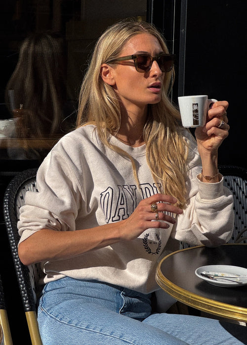 Femme assise à un café portant le sweatshirt beige de la collection capsule Day+ University.