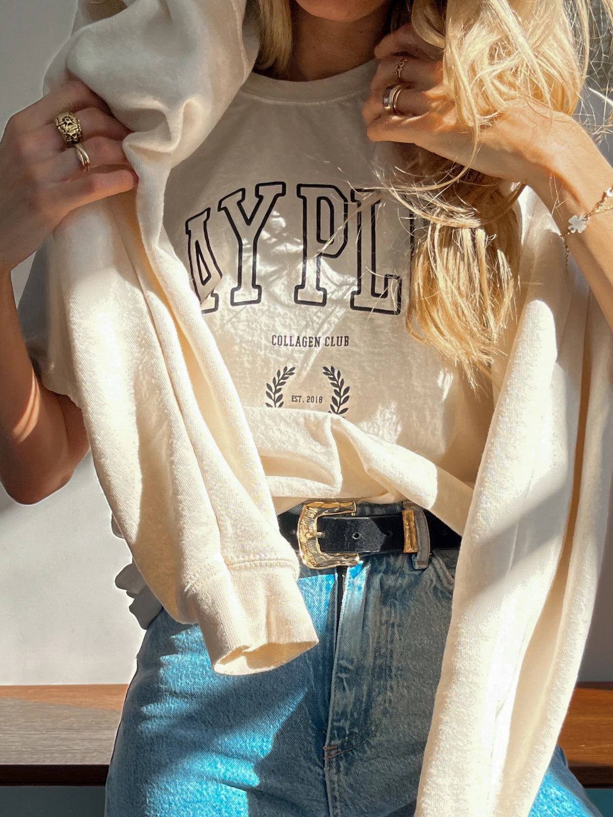 Femme portant le T-shirt beige de la Day+ University avec le sweat de la même collection sur les épaules.