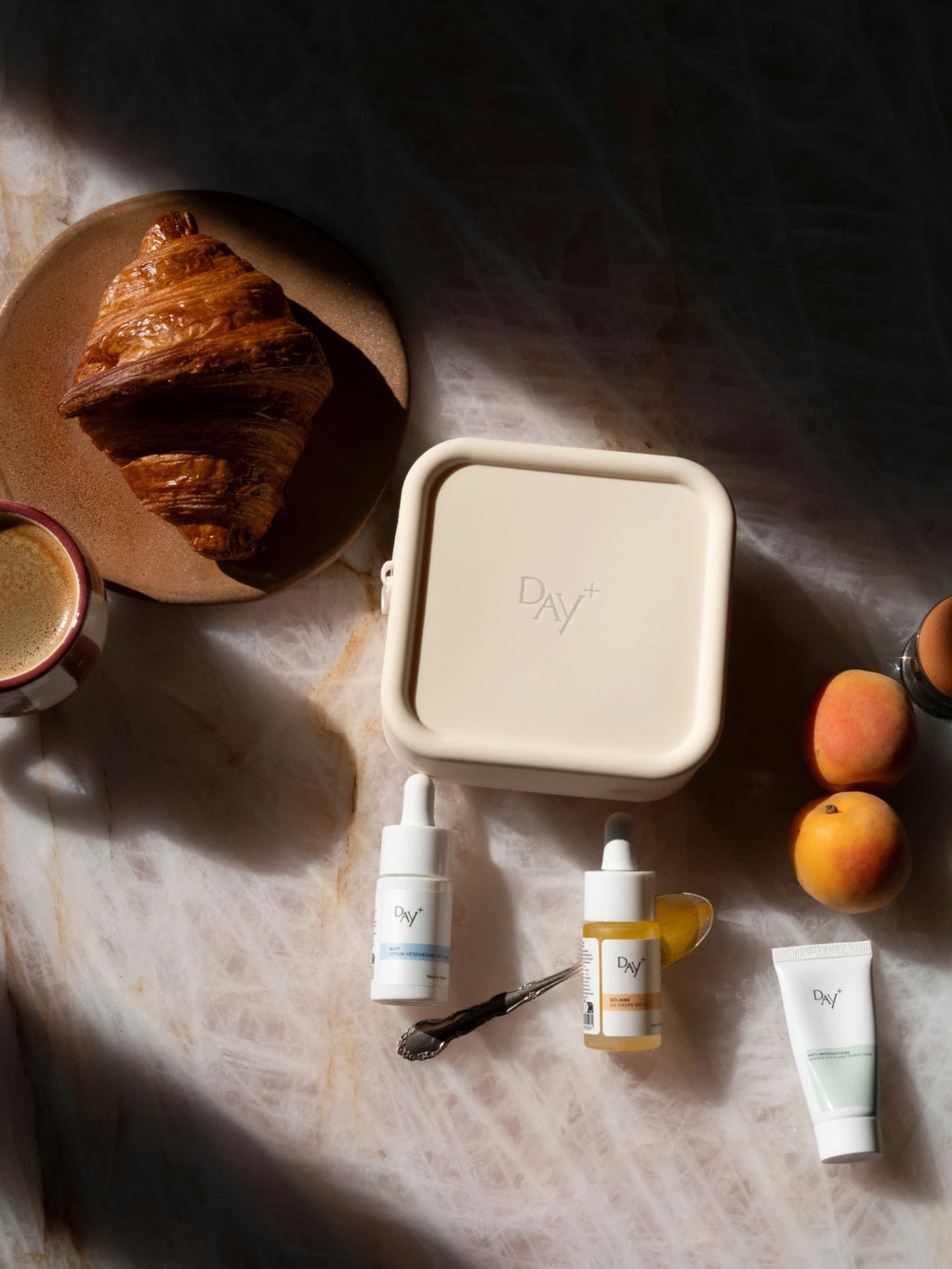 Photo de la trousse beige de voyage Day+ posée sur une table avec 3 produits en format travel, un café, un croissant et des abricots