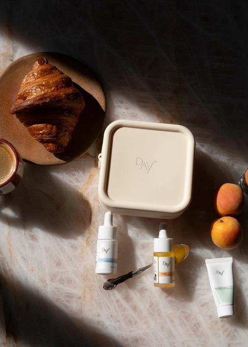 Photo de la trousse beige de voyage Day+ posée sur une table avec 3 produits en format travel, un café, un croissant et des abricots